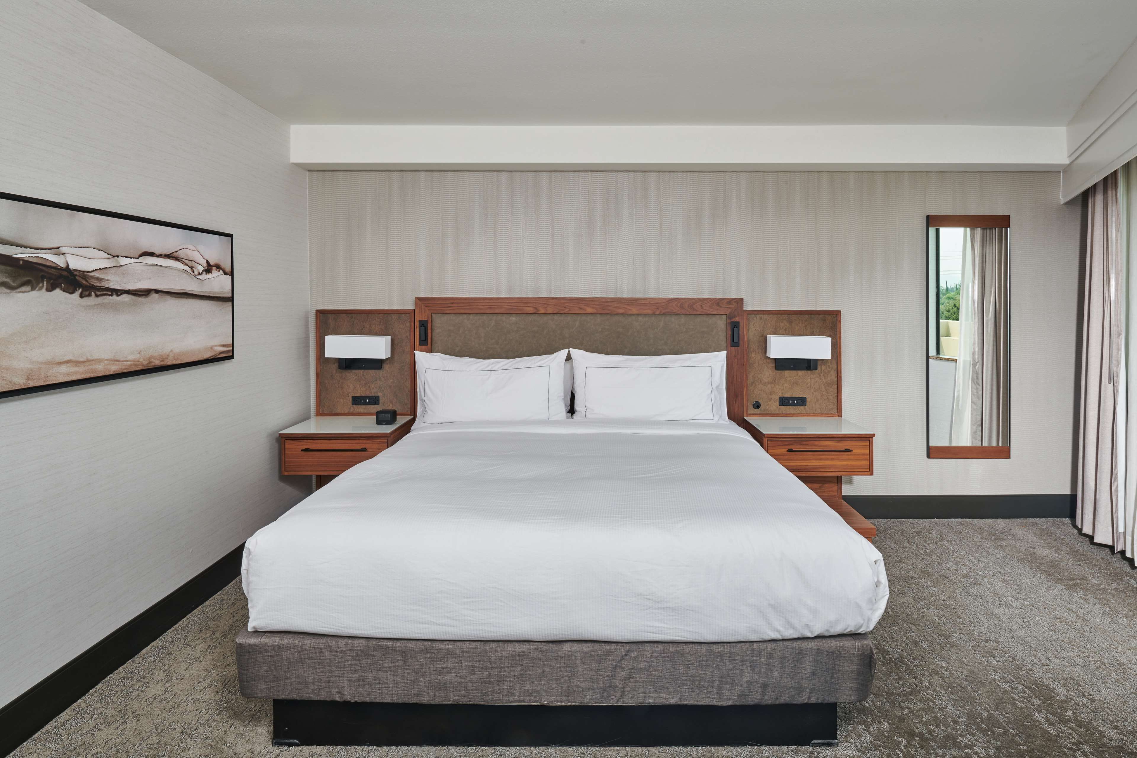 Une chambre d’hôtel avec un grand lit, deux tables de chevet, une petite table, une lampe fixée au mur et une fenêtre avec des rideaux.