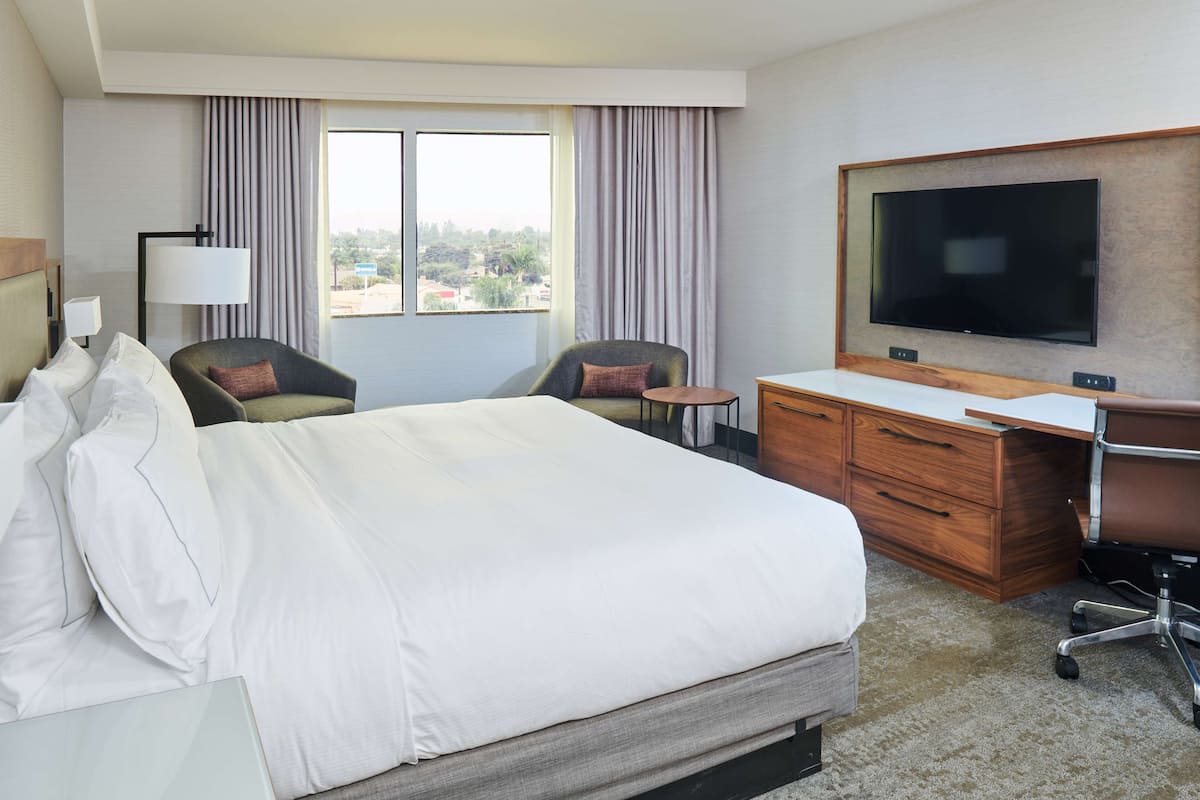 A hotel room with a large bed, two armchairs, a desk, a television, and a window with curtains.
