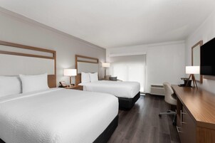 A hotel room with two beds, a desk, a chair, a TV, and a lamp.