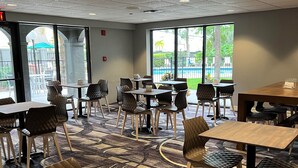 Un restaurant au décor moderne, avec un mélange de chaises en bois et de chaises rembourrées, ainsi que des tables surmontées d’un tapis à motifs. De grandes fenêtres offrent une vue sur l’espace extérieur.