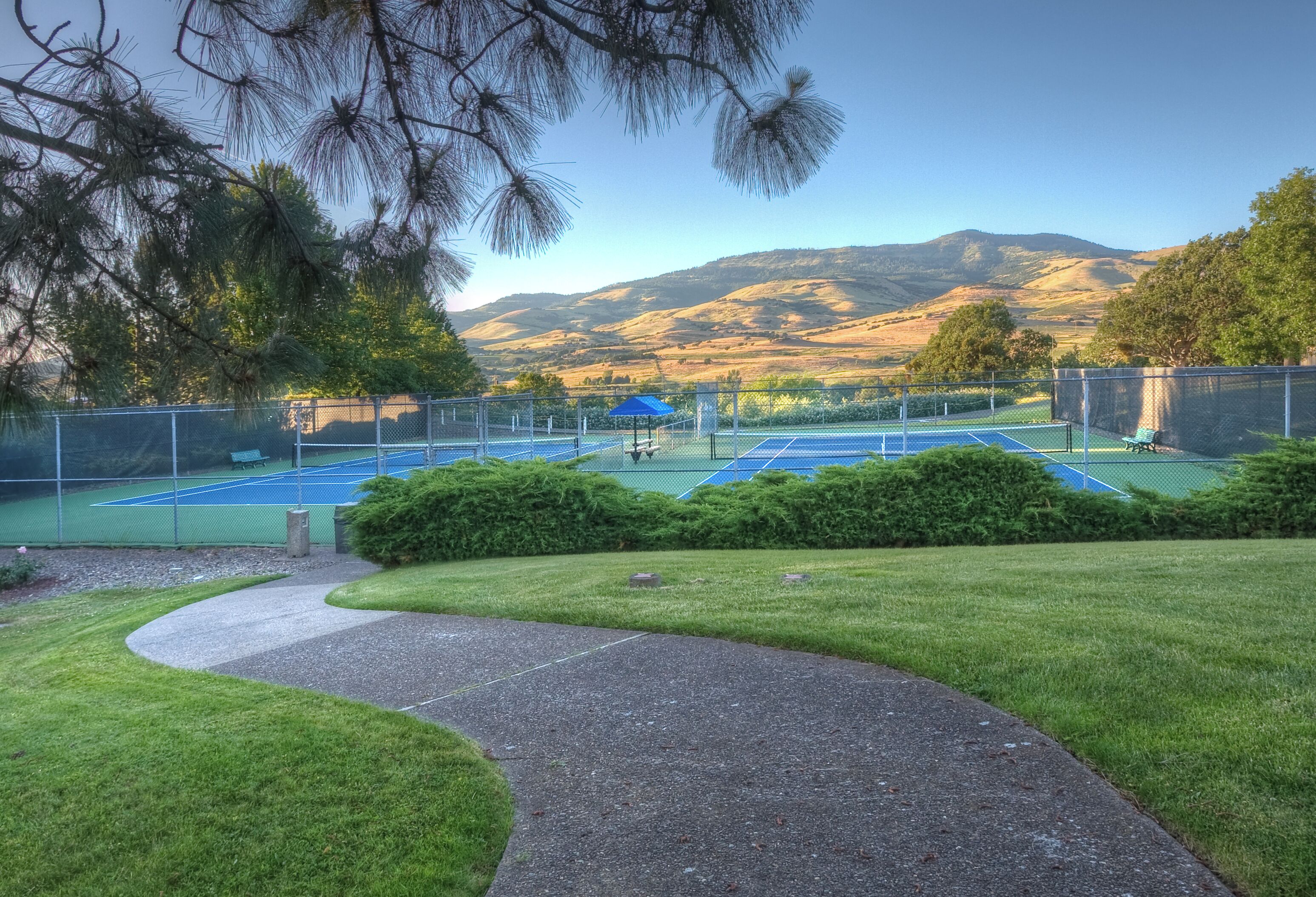 A tennis court with a pathway leading to it, surrounded by greenery and a mountain backdrop.