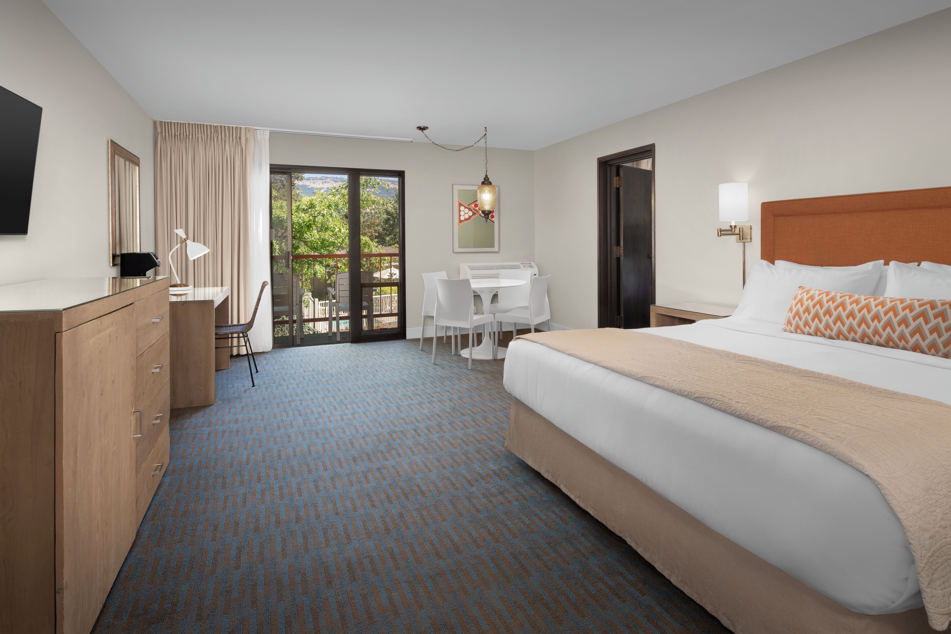 A hotel room with a large bed, a desk with a chair, a wardrobe, a TV mounted on the wall, and a view of the outdoors through a glass door.