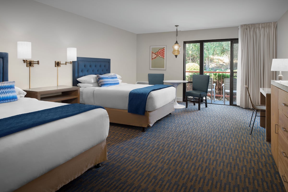 A hotel room with two beds, a desk, and a chair. There is a window with a view of greenery outside.