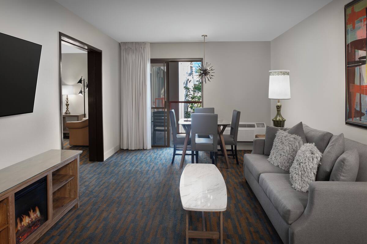 A modern hotel room with a flat-screen TV, a fireplace, a grey sofa, a dining table with chairs, and a large window with a view of the city.