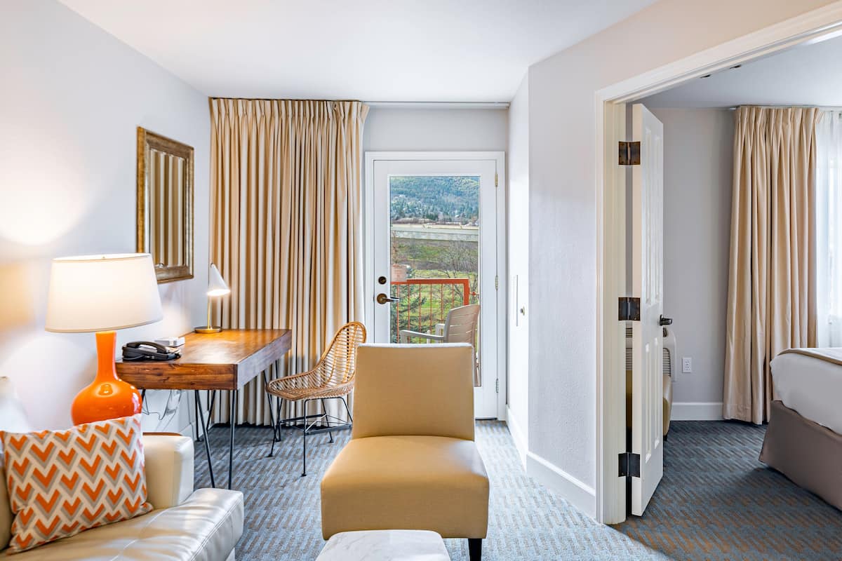 A hotel room with a sofa, a chair, a desk with a lamp, a bed, and a view of the outdoors through a door.
