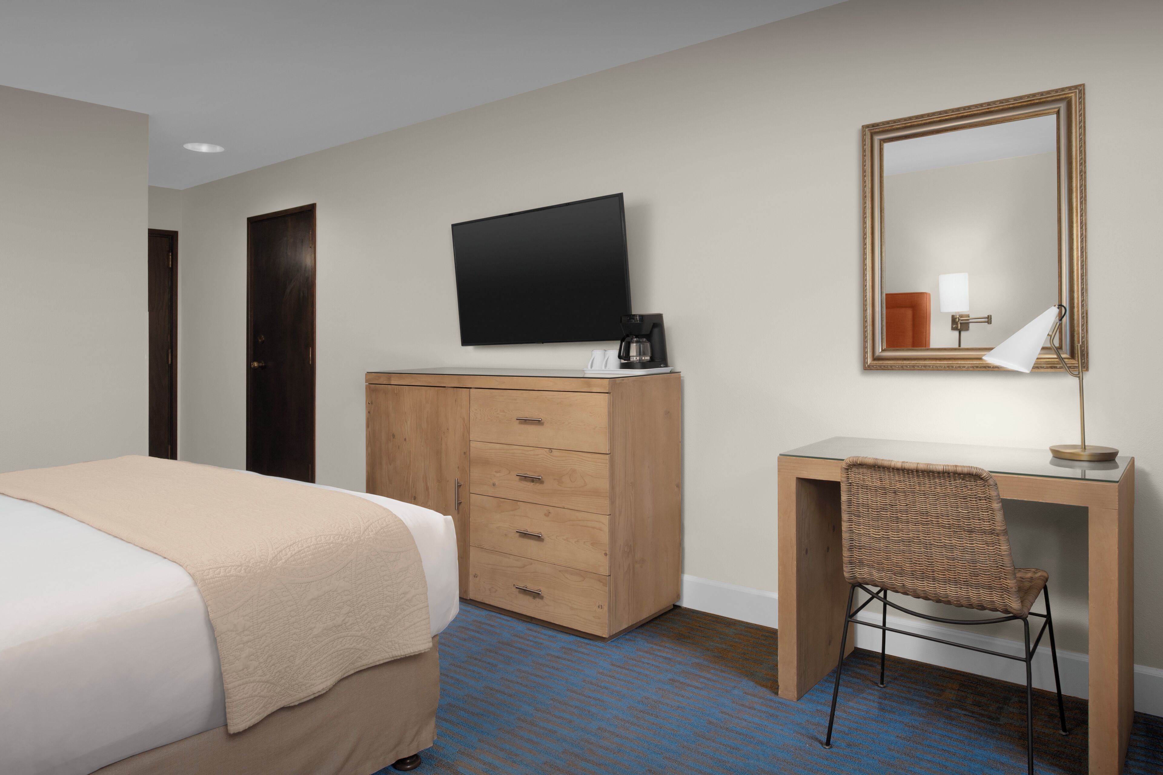 A hotel room with a bed, a wooden desk, a chair, a television, a mirror, and a coffee maker.