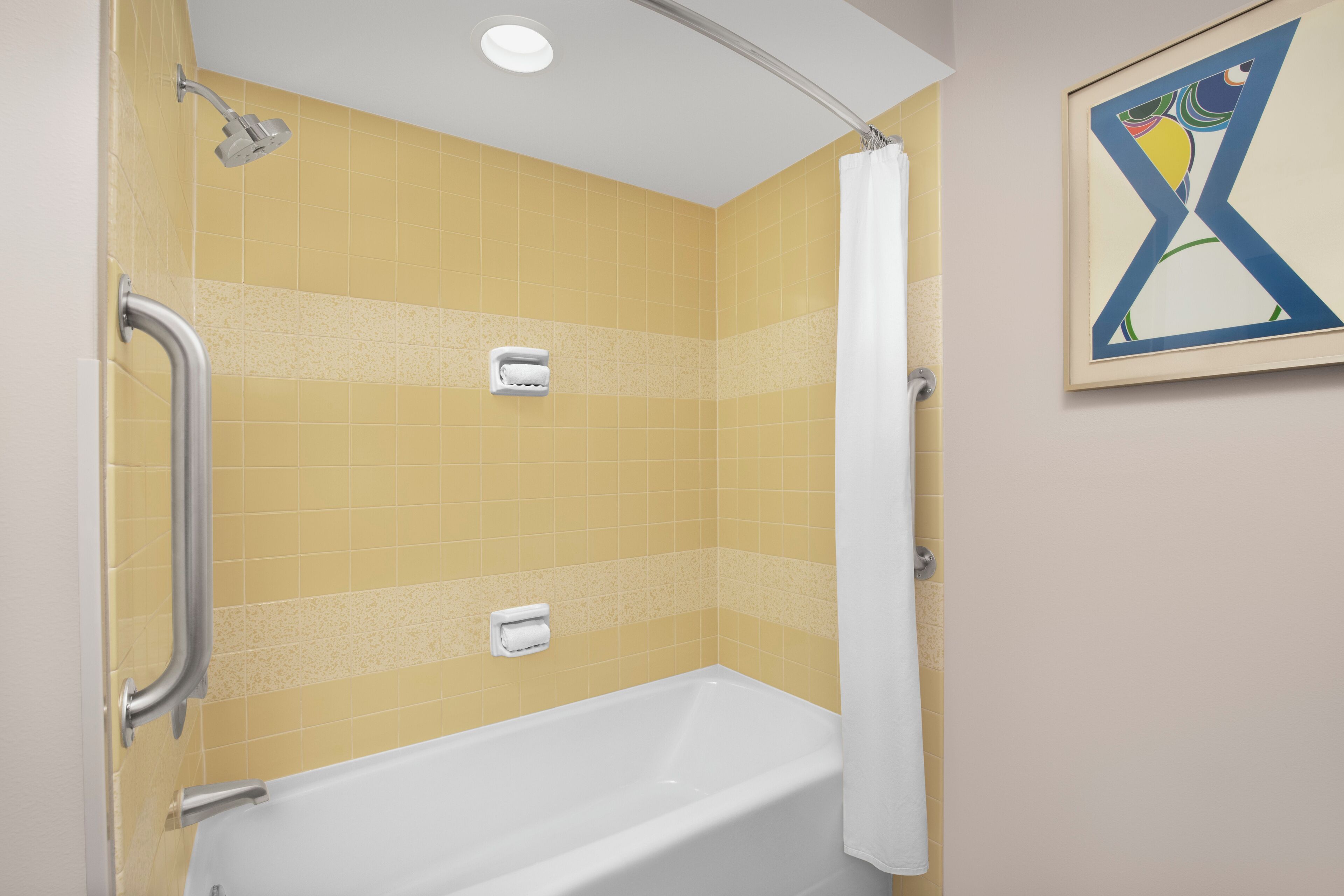A bathroom with a white bathtub, yellow tiled walls, a handrail, a showerhead, and a framed abstract painting.