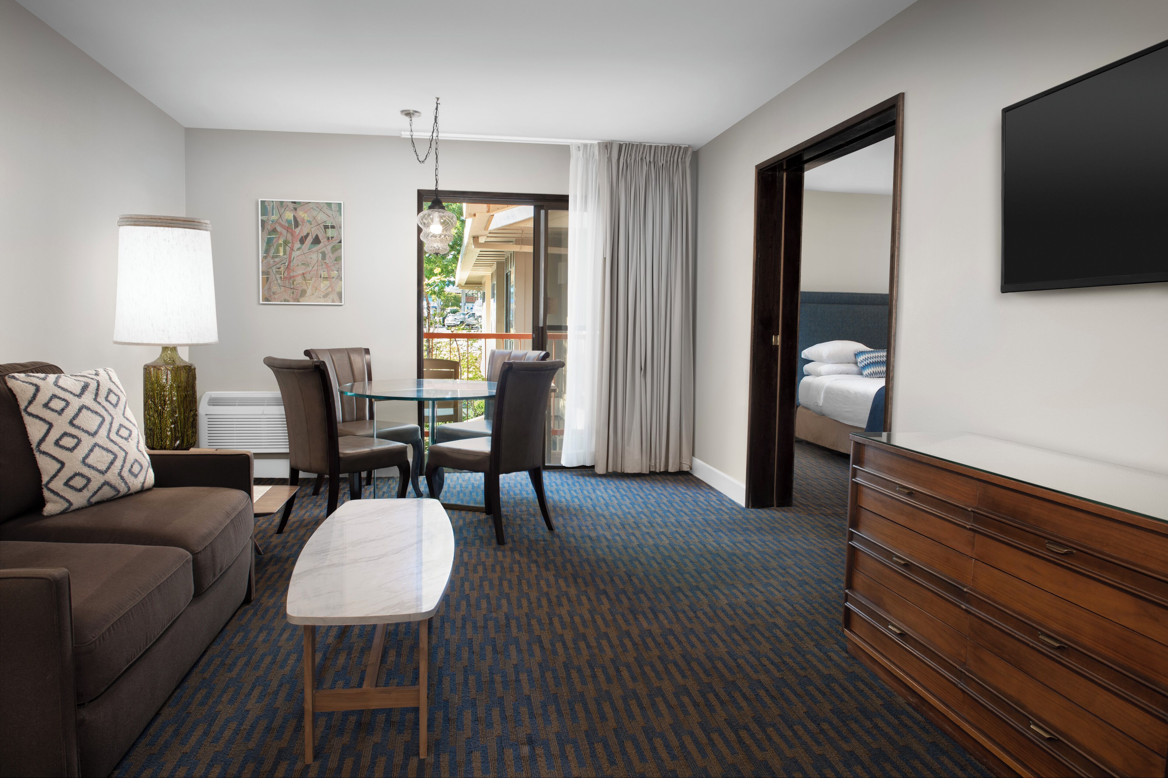 Une chambre d’hôtel avec un canapé, une table, une commode, une chaise, une lampe, un tableau et une vue sur l’extérieur.