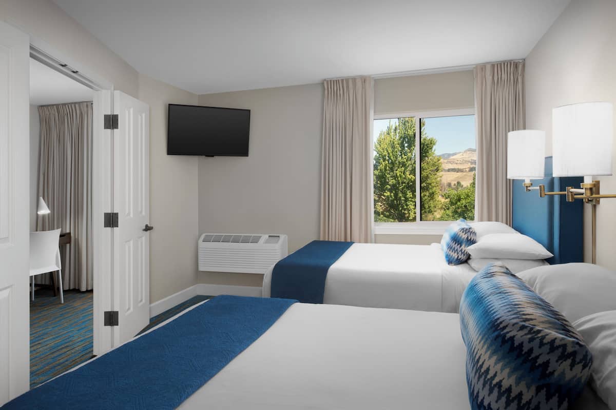 A hotel room with two beds, a TV, an air conditioning unit, and a window with a view of trees and hills.