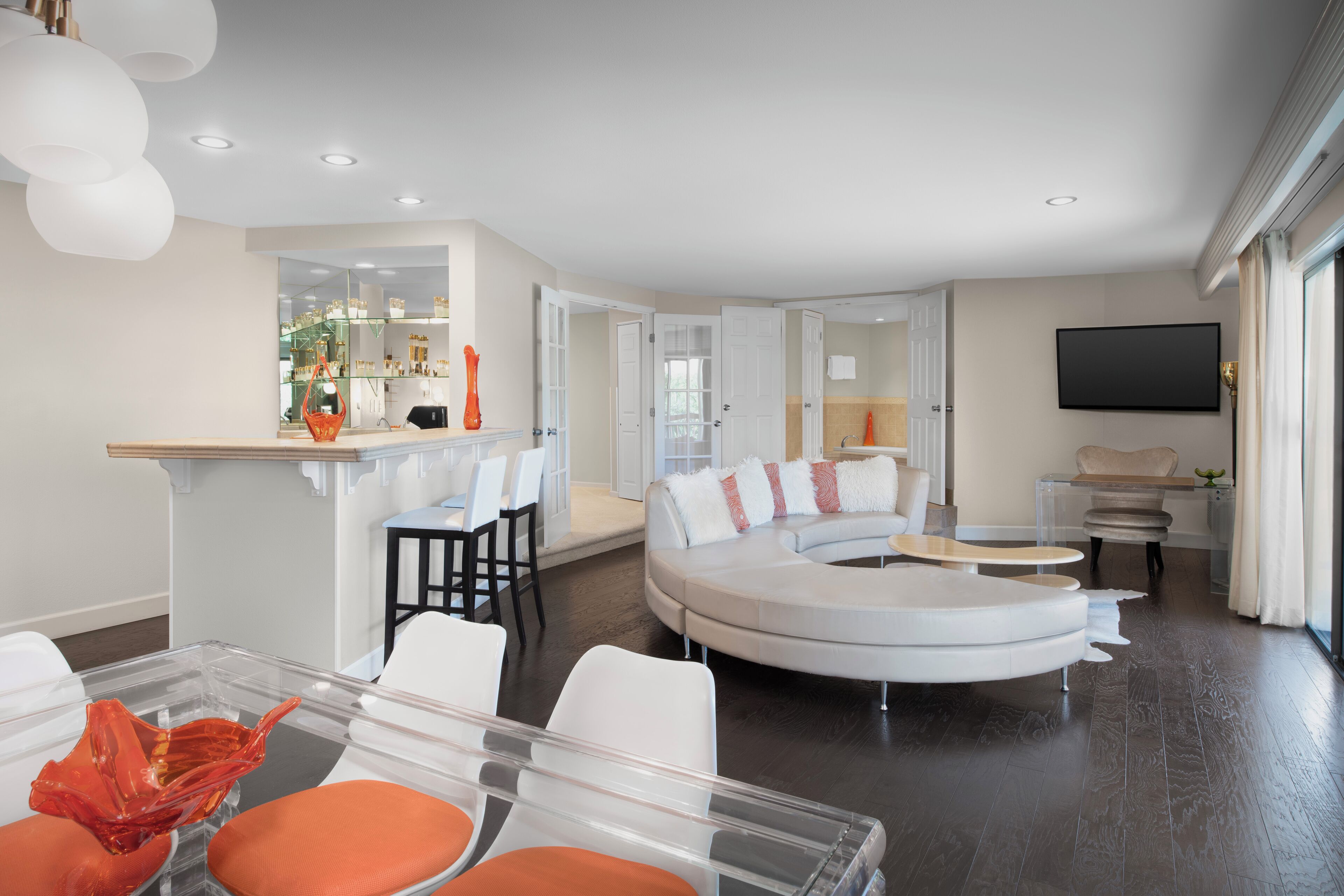 A modern living room with a curved white sofa, a glass coffee table, and a dining area with a clear table and orange chairs.