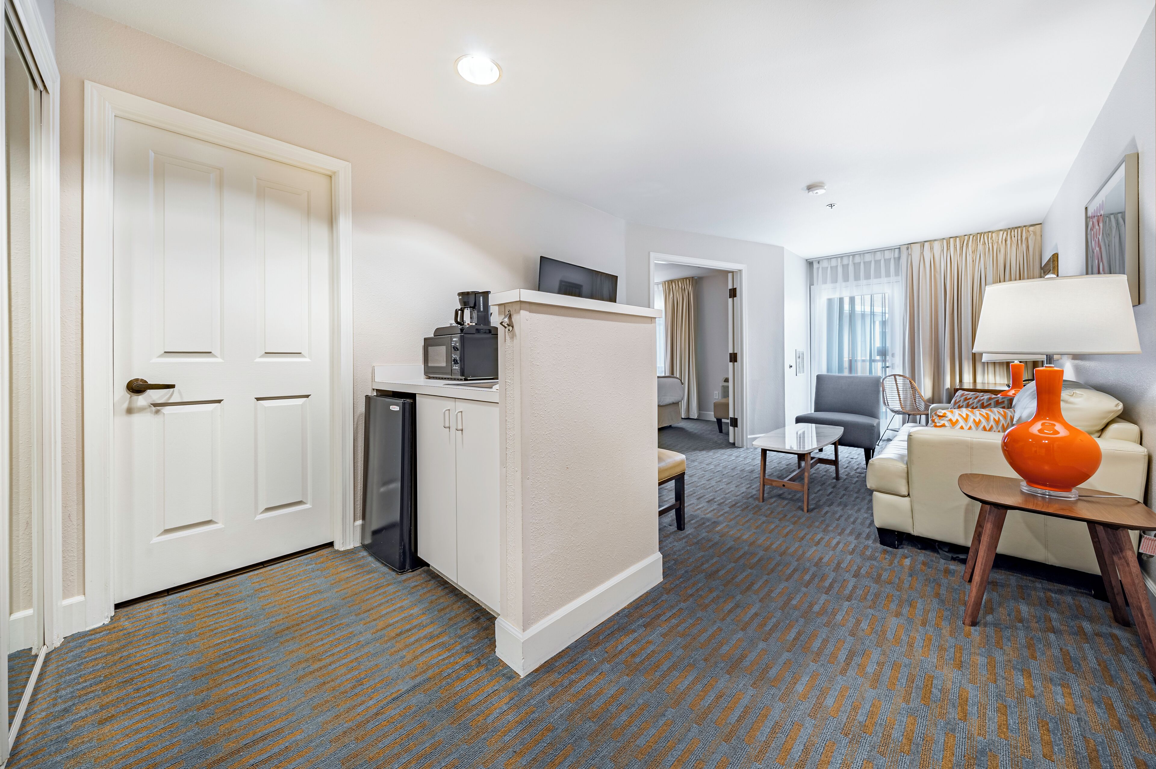 Quarto de hotel com cozinha compacta, sala de estar com sofá e poltrona, mesa de jantar e uma grande janela com cortinas.