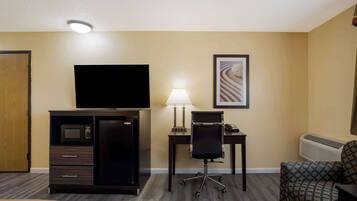 A hotel room with a flat-screen TV, a desk with a lamp, a chair, a patterned sofa, a minibar, and a door.