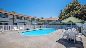 Piscina com espreguiçadeiras e guarda-sóis, cercada por uma vedação branca e um edifício com várias janelas.
