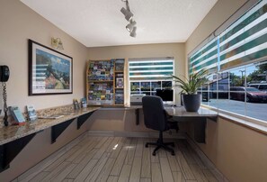 A reception area with a counter, a framed painting, a bookshelf, a desk with a chair, a potted plant, and a large window with a view of parked cars. - Quality Inn & Suites Medford Airport (Medford)