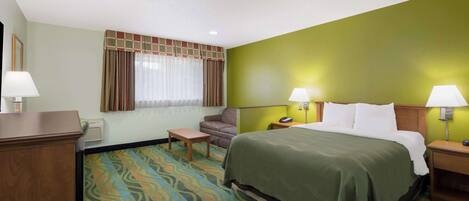 Une chambre d’hôtel comprenant un lit, deux tables de chevet avec des lampes, une chaise, un bureau, une fenêtre avec des rideaux et une moquette à motifs.