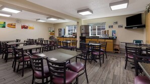 Una cafetería con varias mesas y sillas, una zona de cocina y un televisor en la pared.