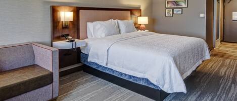 A hotel room with a large bed, a bedside table with a lamp, a chair, and a wall with framed pictures.