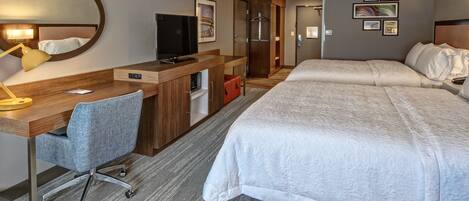 A hotel room with a bed, desk, chair, TV, and artwork on the wall.
