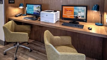 Uma mesa de recepção com dois monitores de computador, uma impressora e um teclado. Há duas cadeiras de escritório e um abajur.
