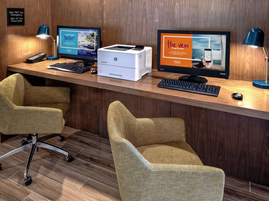 Un mostrador de recepción con dos monitores de computadora, una impresora y un teclado. Hay dos sillas de oficina y una lámpara de escritorio.