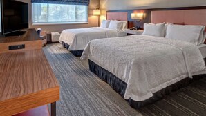 A hotel room with two beds, a TV, a desk, and a window with a view of trees.