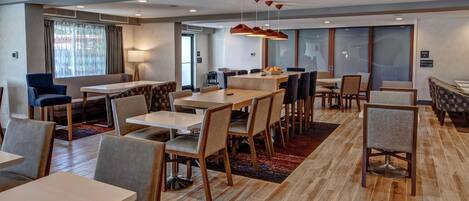 A modern hotel dining area with multiple tables and chairs, a large window with curtains, and a red pendant light.
