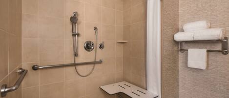 A bathroom with beige tiles, a shower with a handheld showerhead, a grab bar, a towel rack with towels, and a shower seat.
