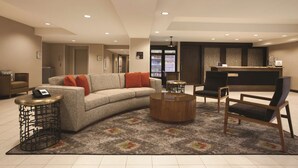 A modern living room with a sofa, coffee table, and chairs. There is a reception desk and a refrigerator in the background. - Homewood Suites by Hilton Tucson/St. Philip's Plaza Univ (Tucson)