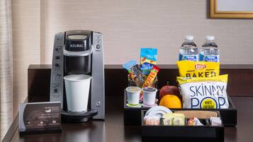 A hotel room's breakfast setup with a Keurig coffee maker, a coffee cup, and a tray of snacks including Lay's Baked Skinny Pop, water bottles, and a box of coffee creamer.