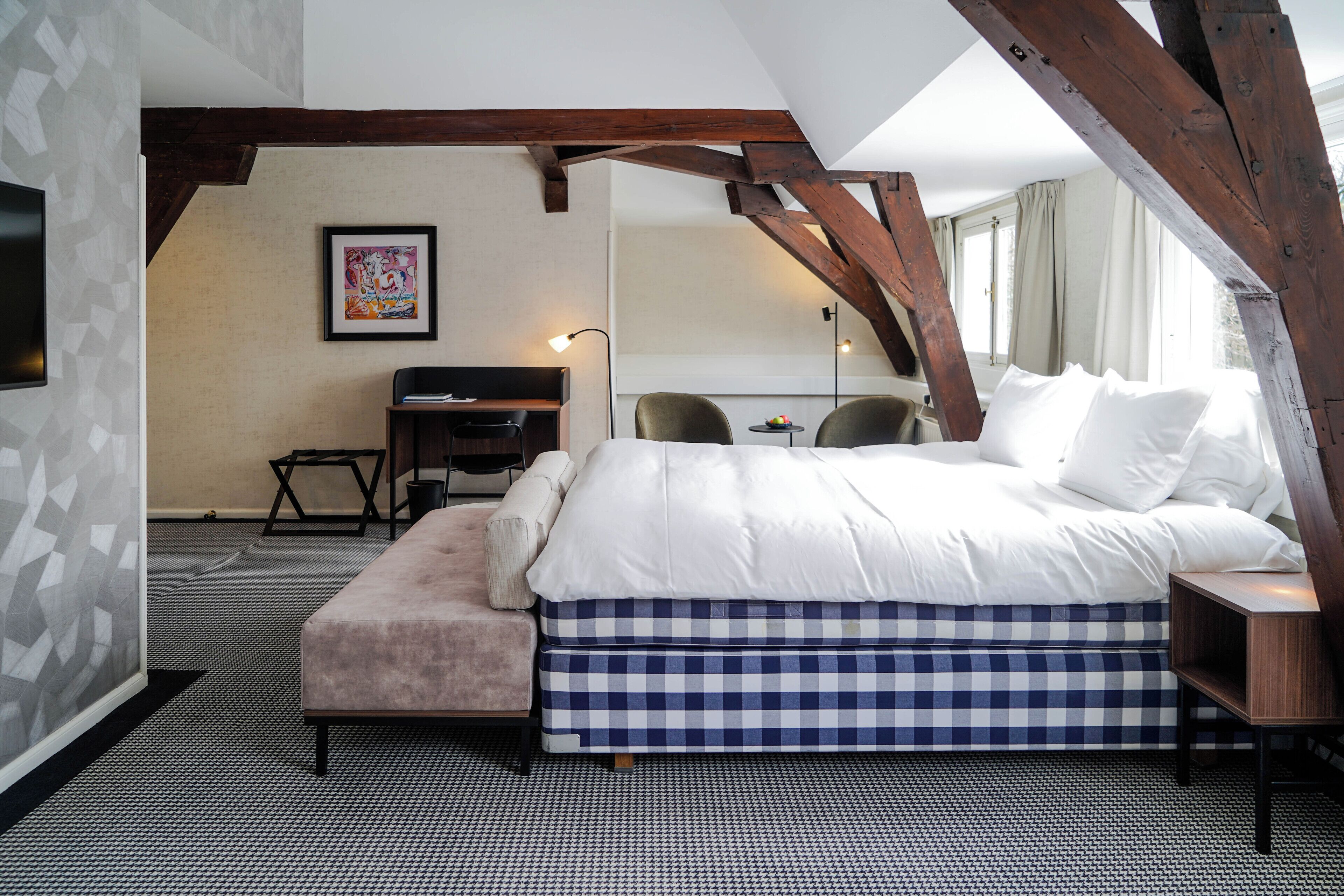 Un dormitorio con una cama grande, un escritorio y una silla. Hay una ventana con cortinas, un cuadro en la pared y una alfombra estampada.
