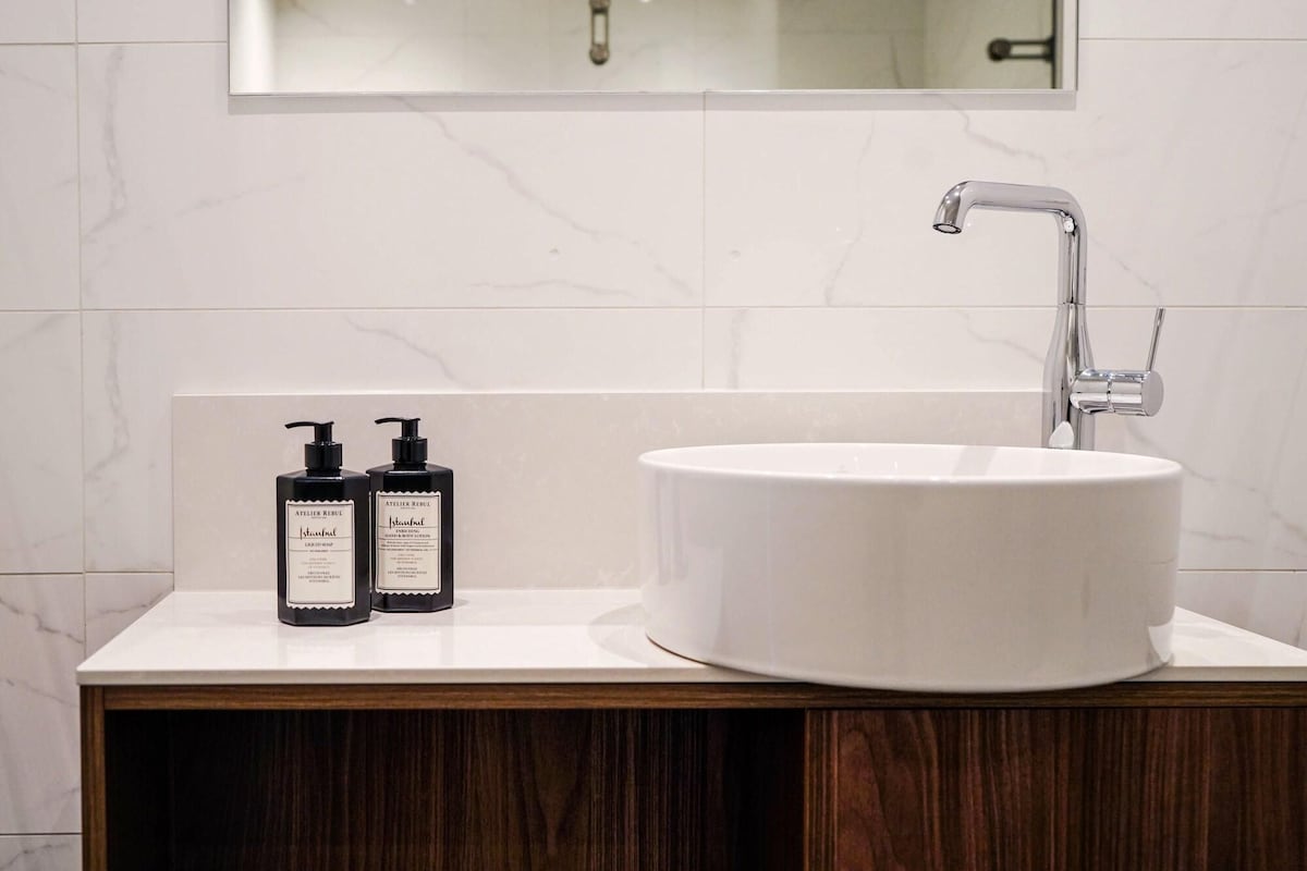 Een badkamer met een witte marmeren wastafel, een moderne kraan en twee flesjes lotion op een houten wastafelmeubel.