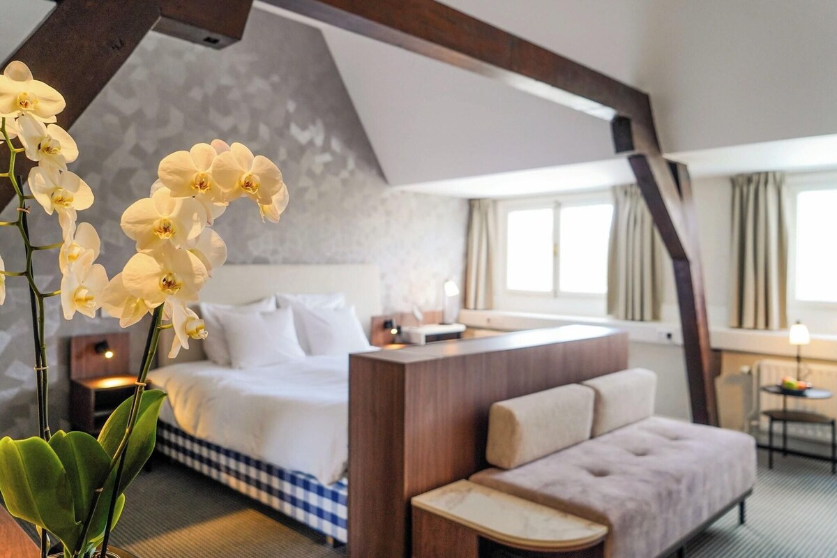 Een hotelkamer met een bed, een bank, een kleine tafel en een bureau. Er is een raam met gordijnen en een plant met witte bloemen.