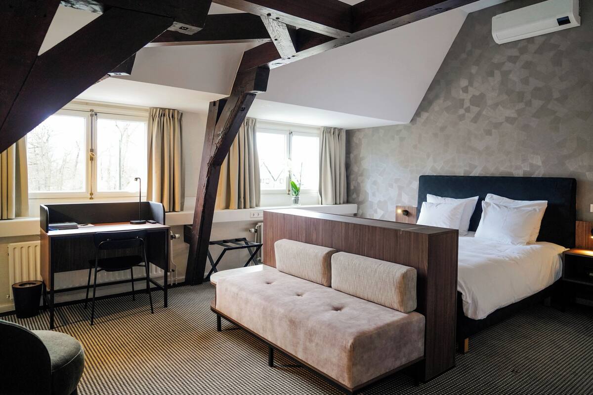 Een moderne hotelkamer met een hoge plafond, een bureau met stoel, een bed met witte lakens, en een klein zithoekje.