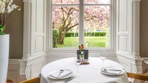 En spiseplads med et rundt bord dækket til to, en vase med blomster og et vindue med udsigt til en have.