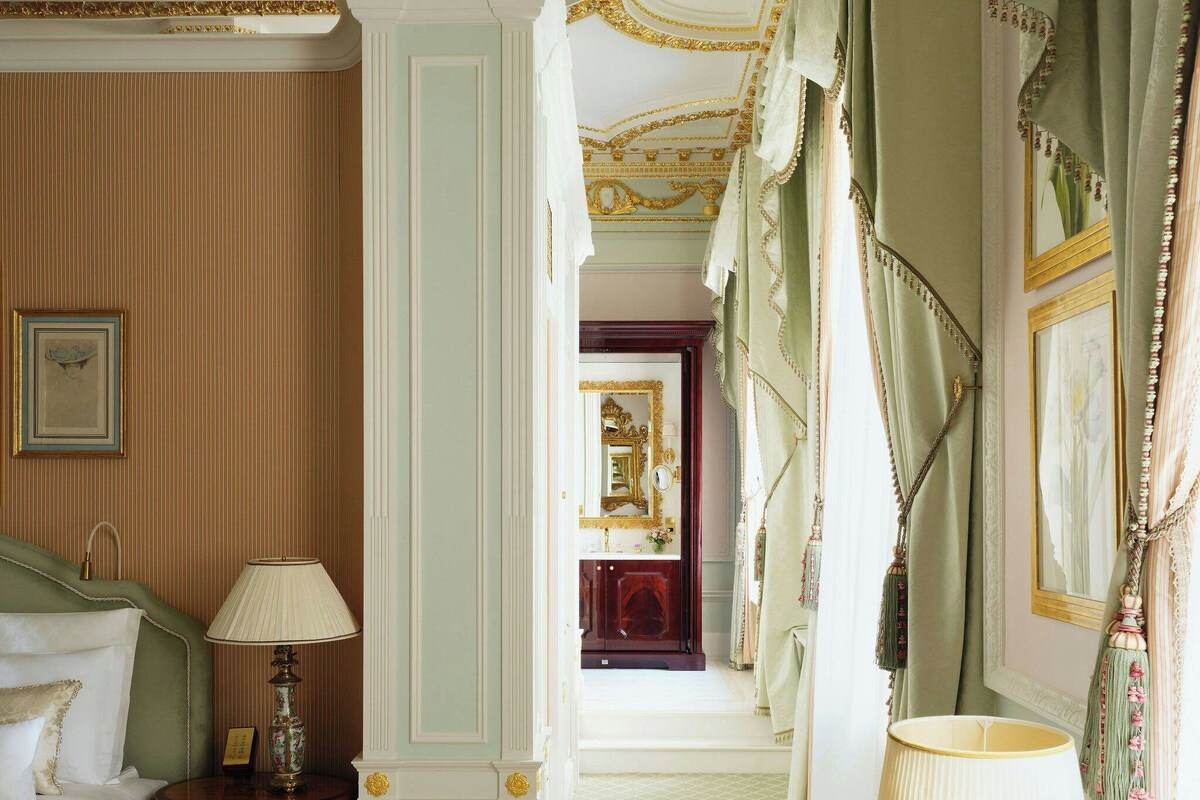 A room with a bed, a chair, a table with a vase of flowers, and a mirror.