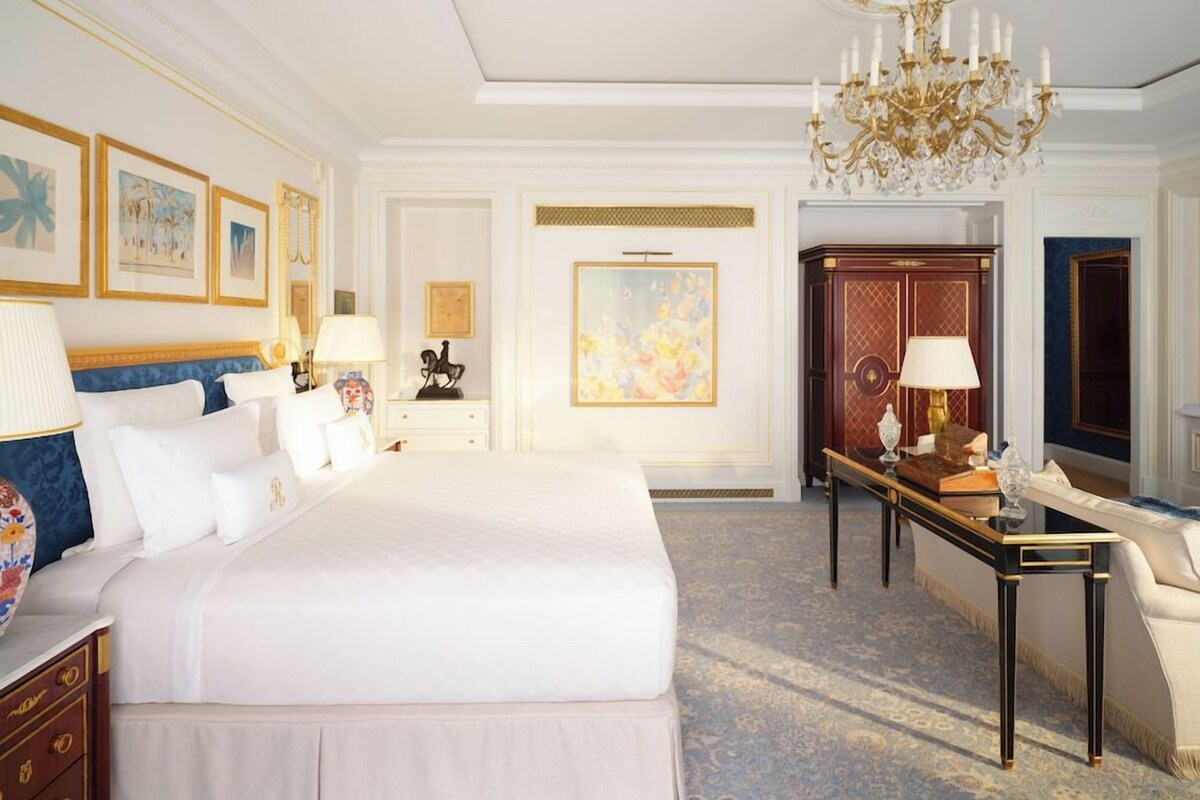 A hotel room with a large bed, a desk, a chair, a chandelier, and framed artwork on the walls.