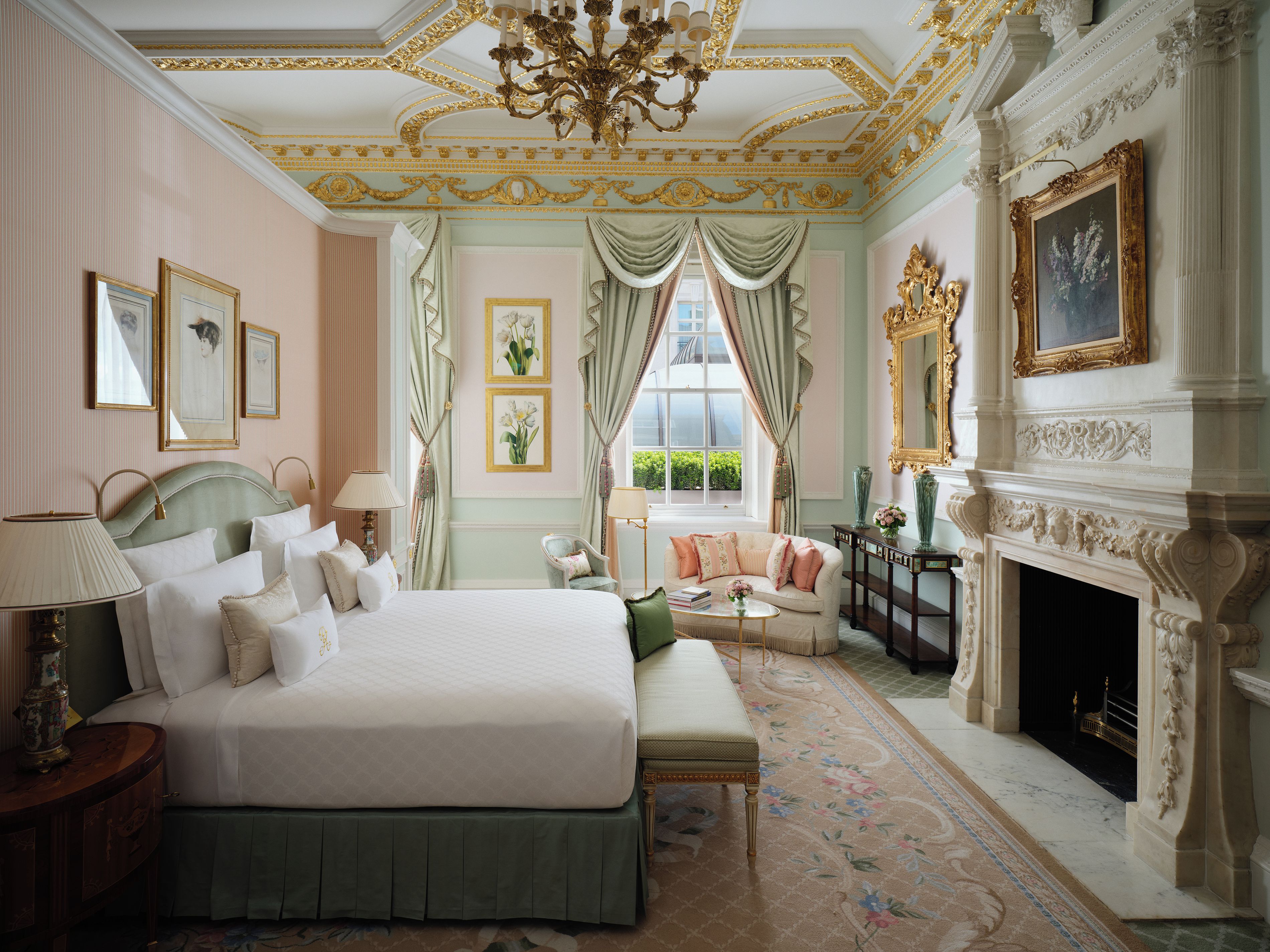 A classic hotel room with a large bed, a sitting area with a sofa and armchairs, a fireplace, and a window with curtains.