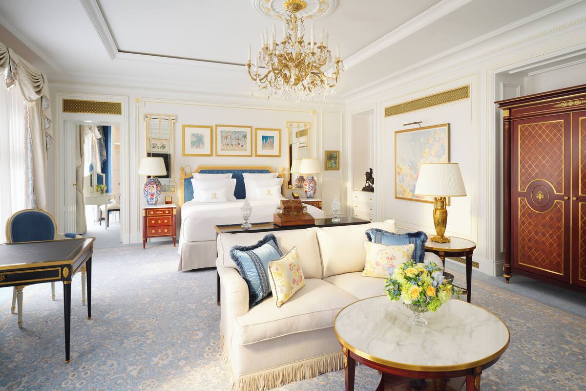 A luxurious hotel room with a large bed, a sofa, a dining table, and a chandelier.