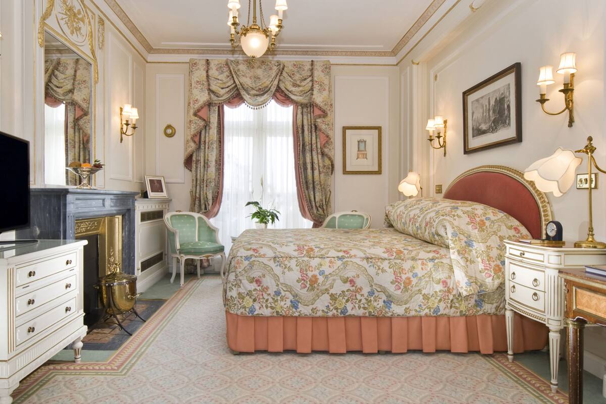 A bedroom with a bed, a dresser, a vanity, a chair, a desk, a television, and a chandelier.