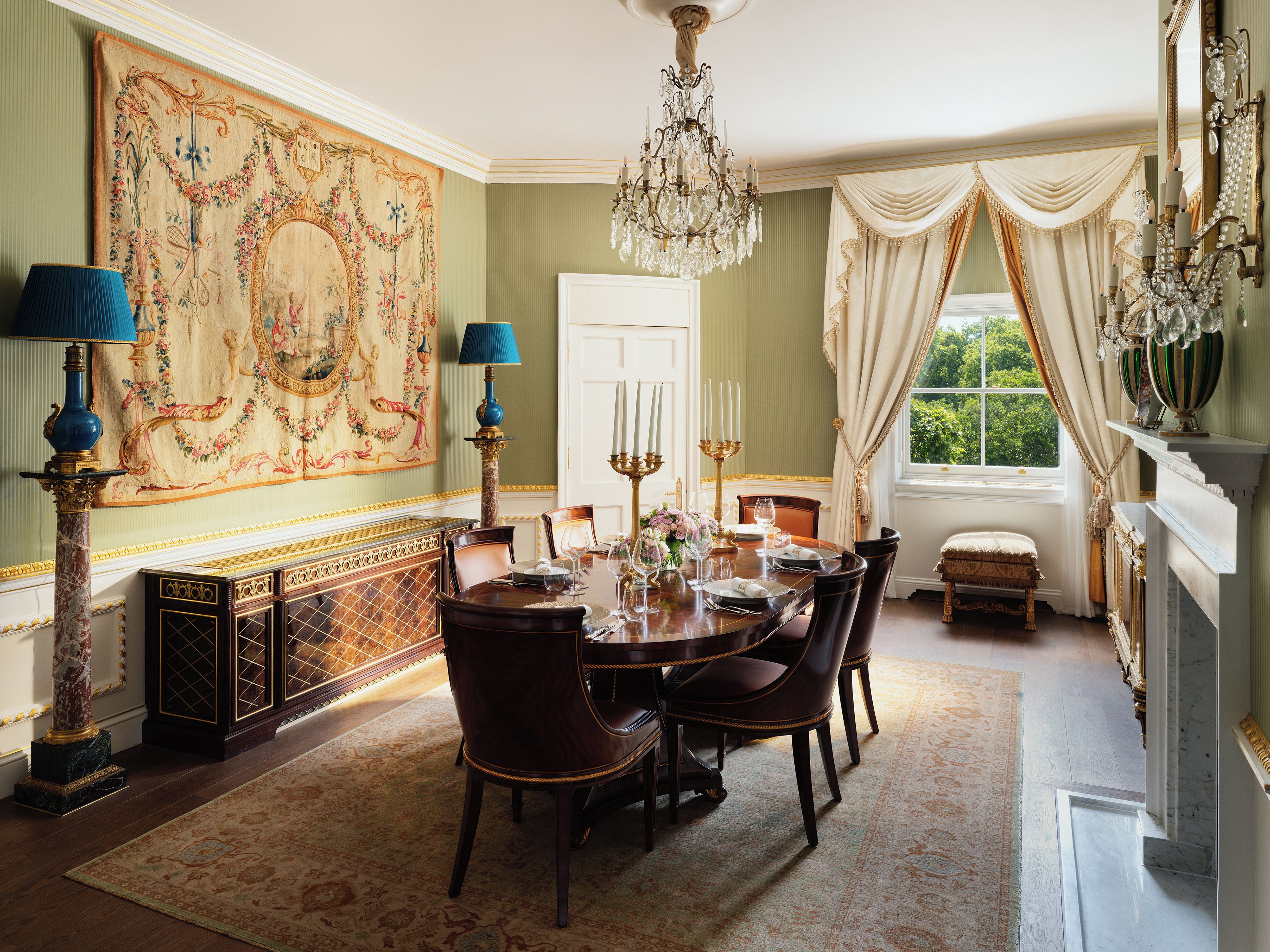 A formal dining room with a chandelier, ornate wall tapestry, and a view of greenery outside.