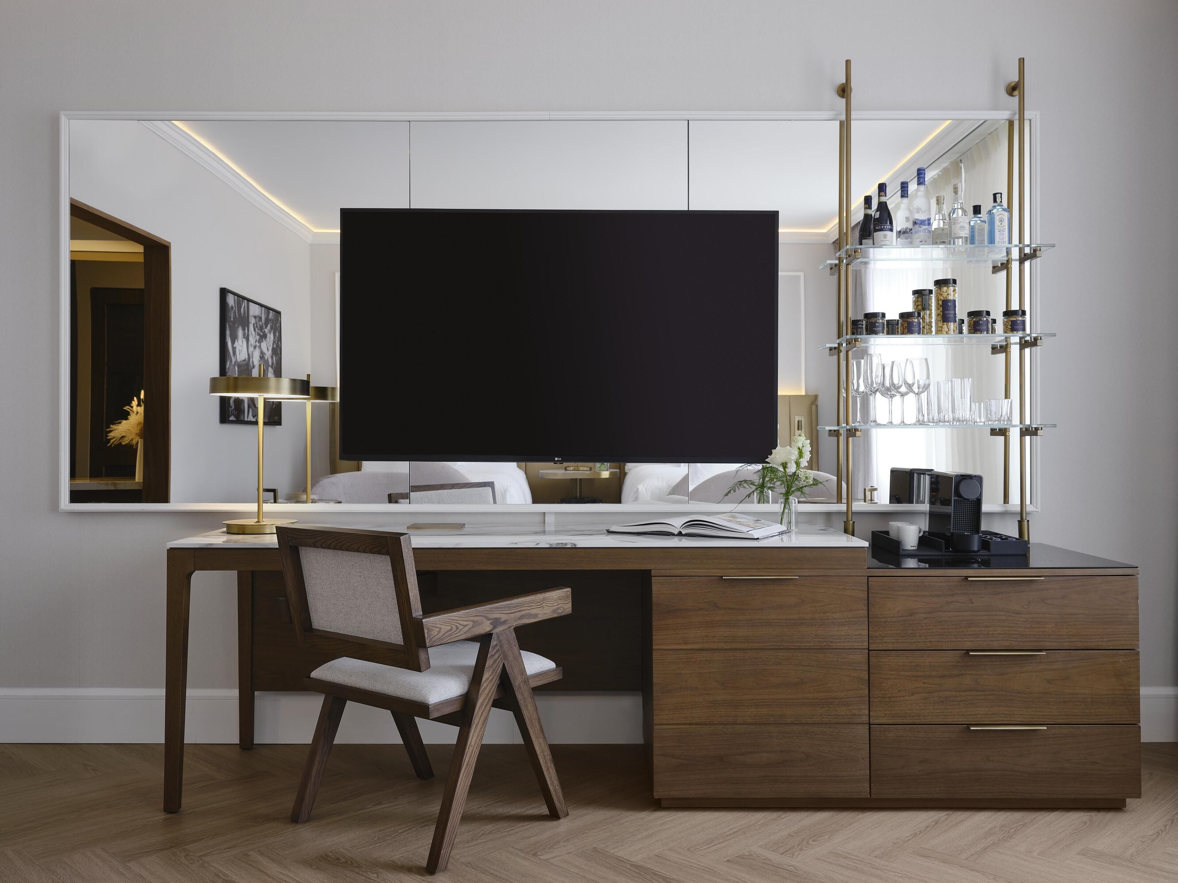 A modern hotel room with a large desk, a chair, a bookshelf, and a large mirror.