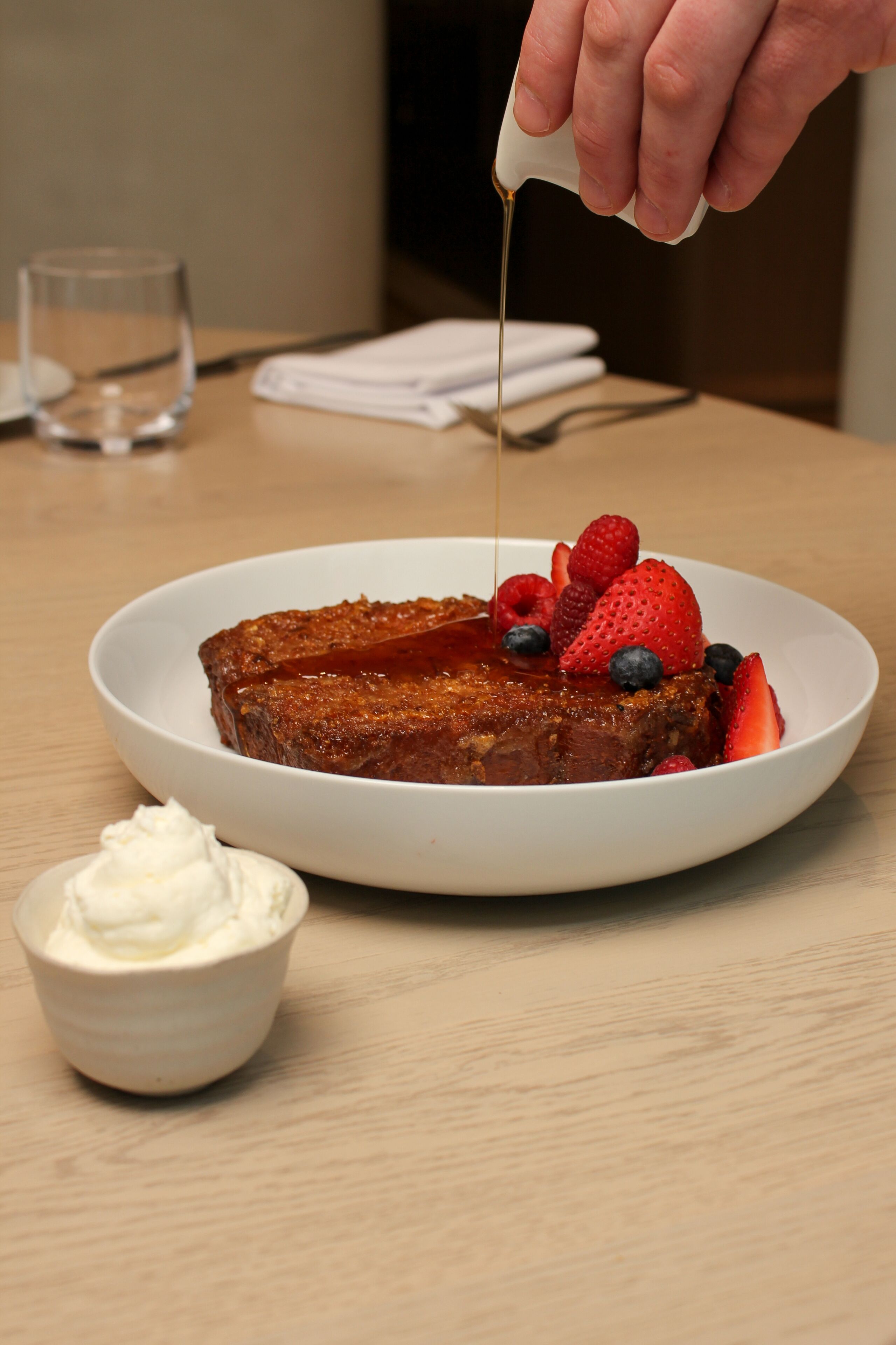 Une assiette de pain perdu garnie de fruits rouges et nappée de sirop, un verre d’eau et un petit bol de crème fouettée, le tout posé sur une table en bois.