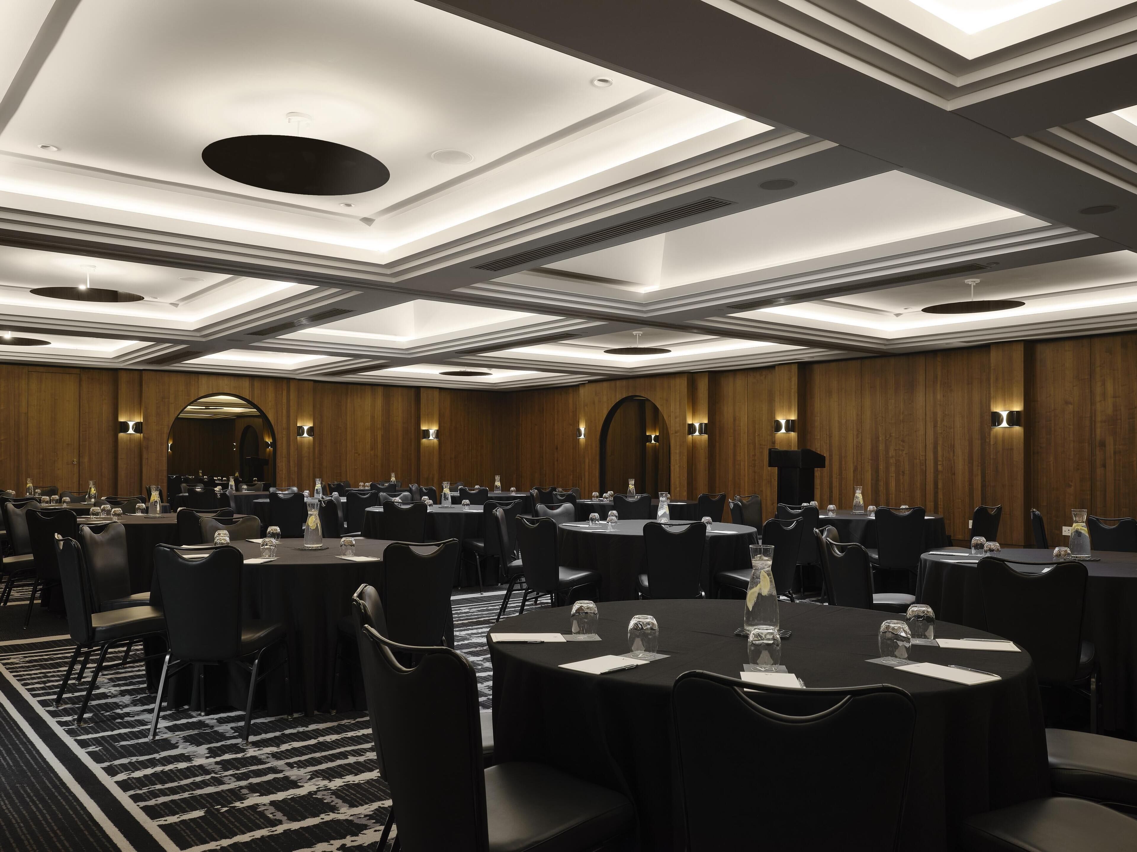 A conference room with multiple round tables, black chairs, and wooden panel walls.