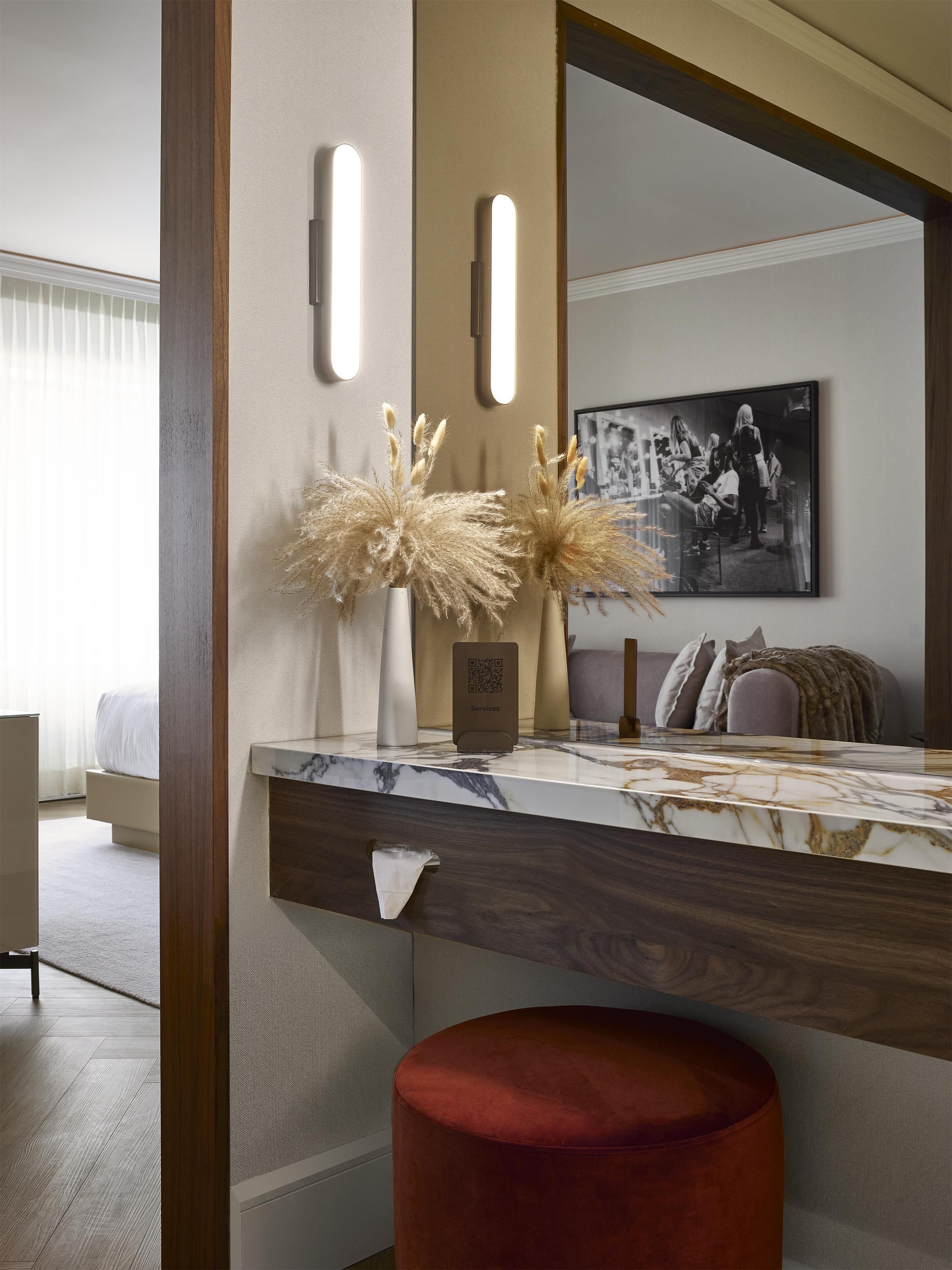 A modern hotel room with a marble countertop, a red ottoman, pampas grass decorations, a framed black and white photograph, and a QR code sign.