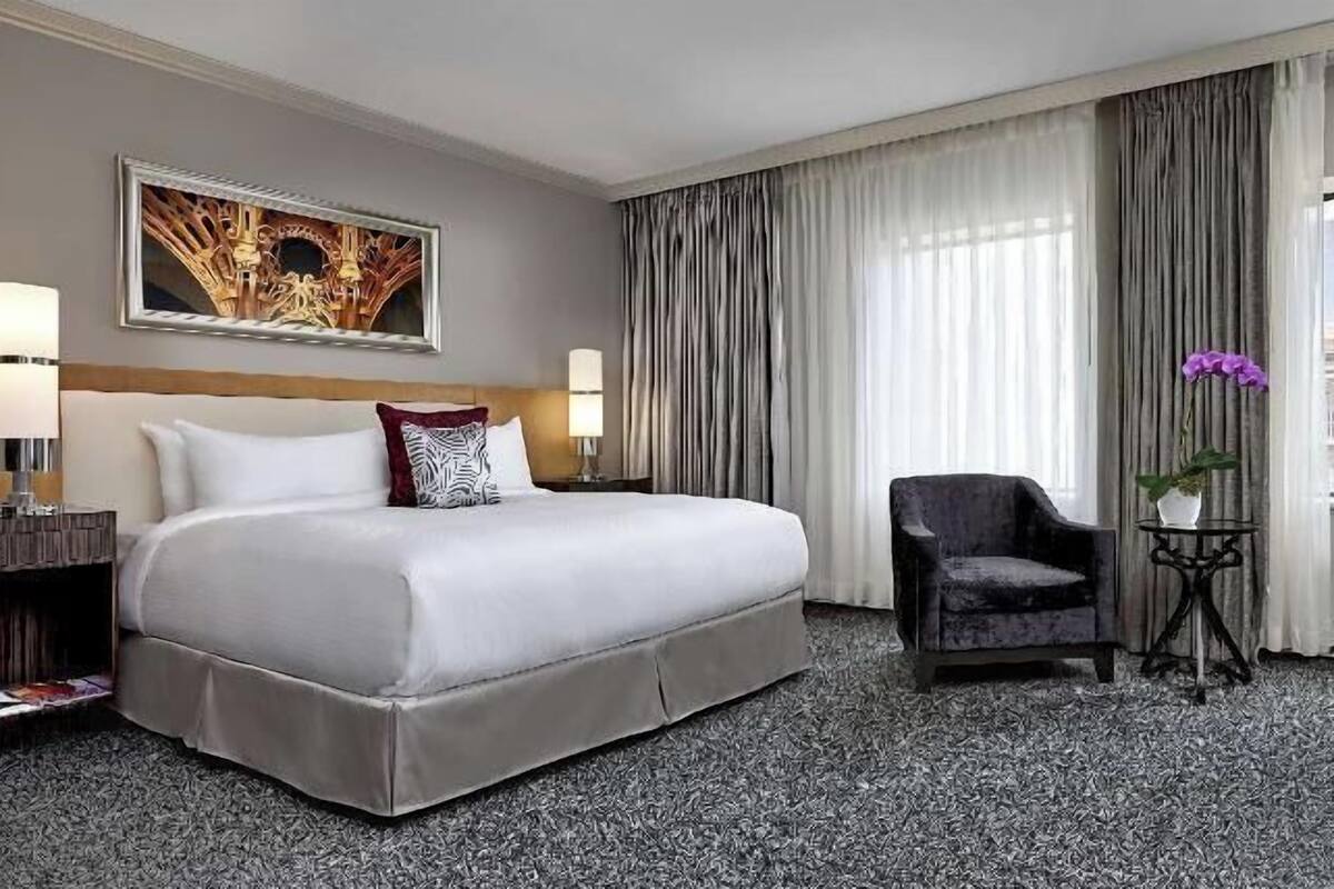 A hotel room with a large bed, a chair, a small table with a vase, and a city view through the window.