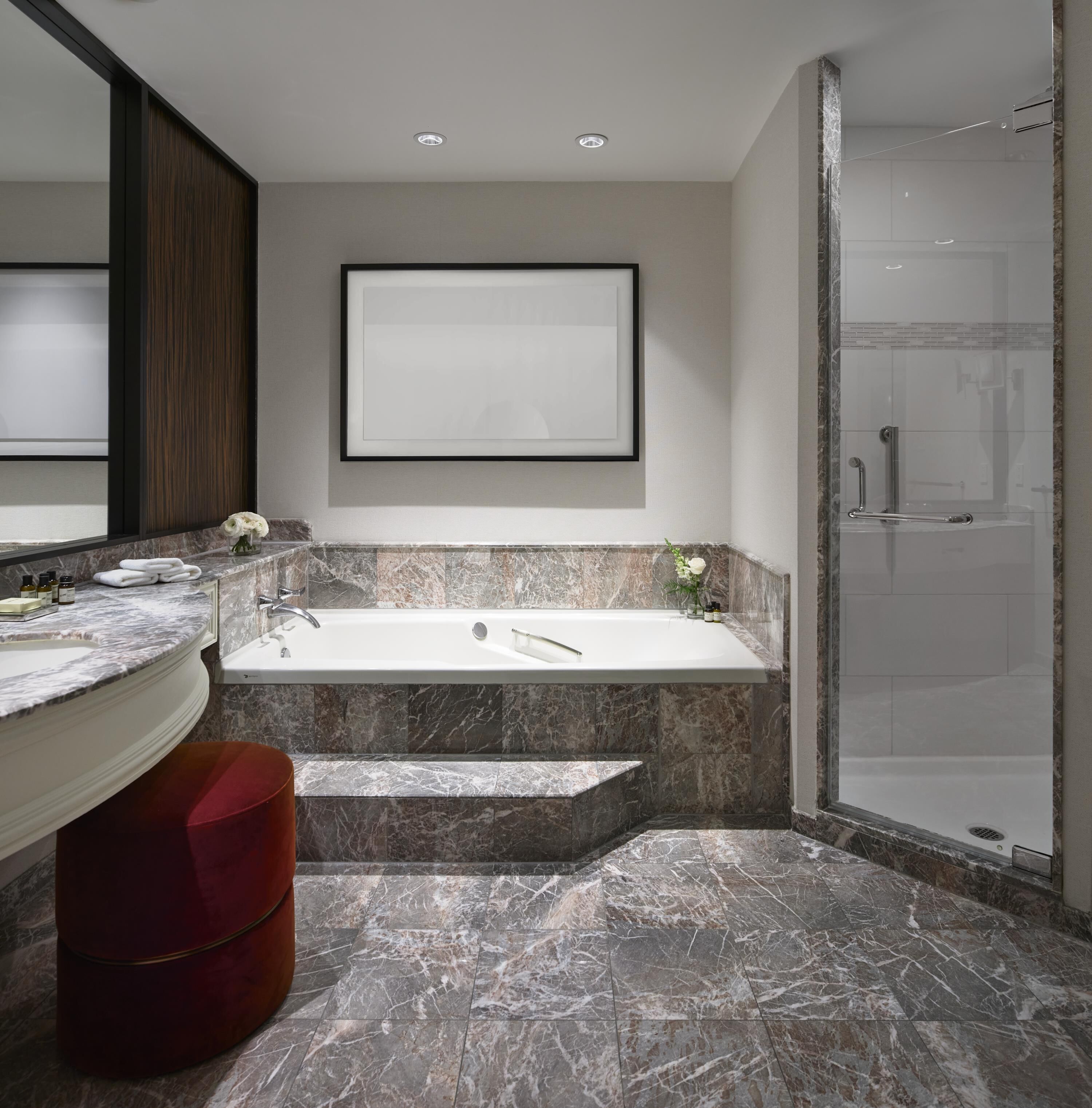 A modern bathroom with a marble sink, a large mirror, a framed artwork, a shower area, and a red stool.