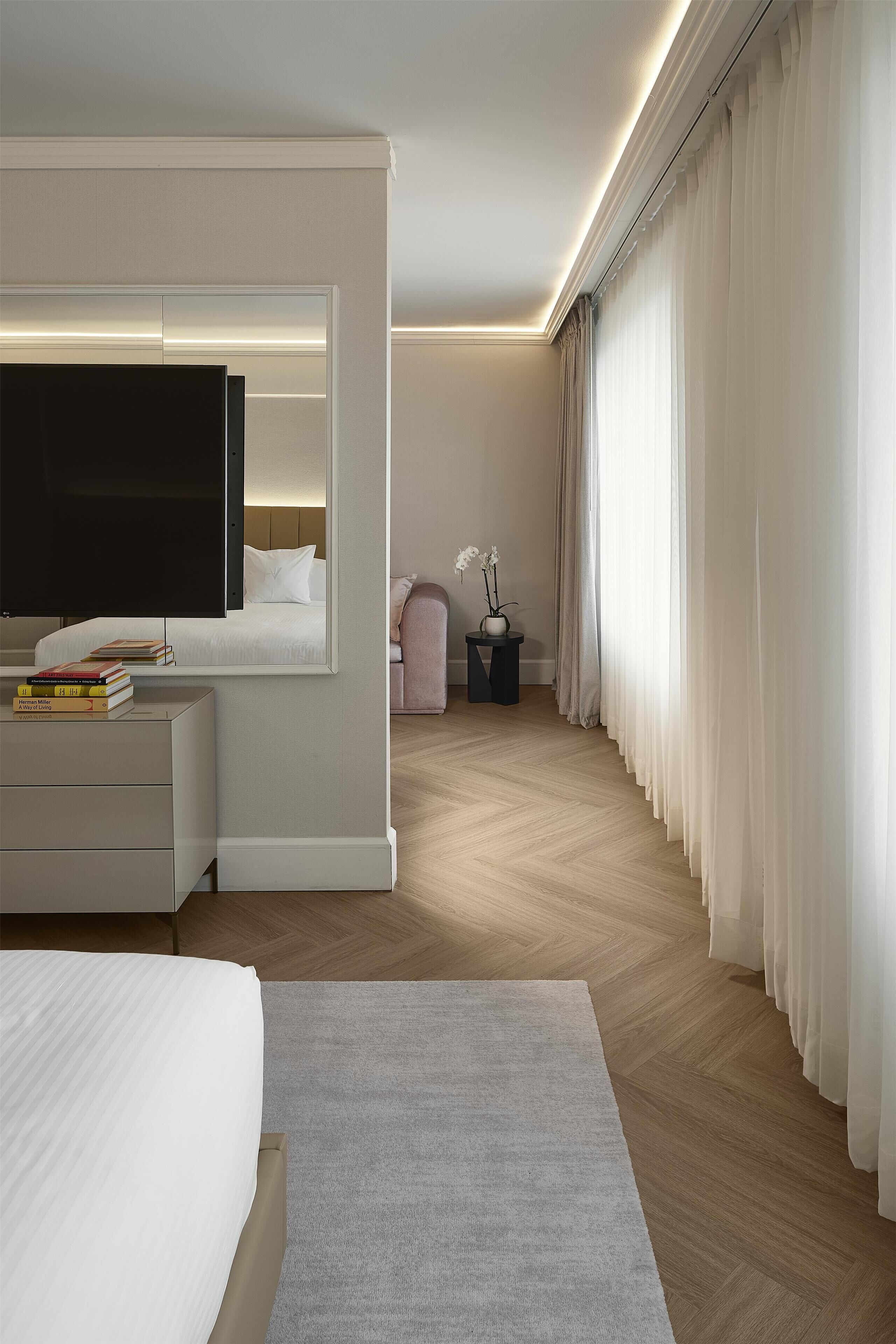 Quarto de hotel moderno com uma cama grande, televisão de tela plana, mesa de cabeceira com livros, mesa lateral com um vaso e cortinas translúcidas.