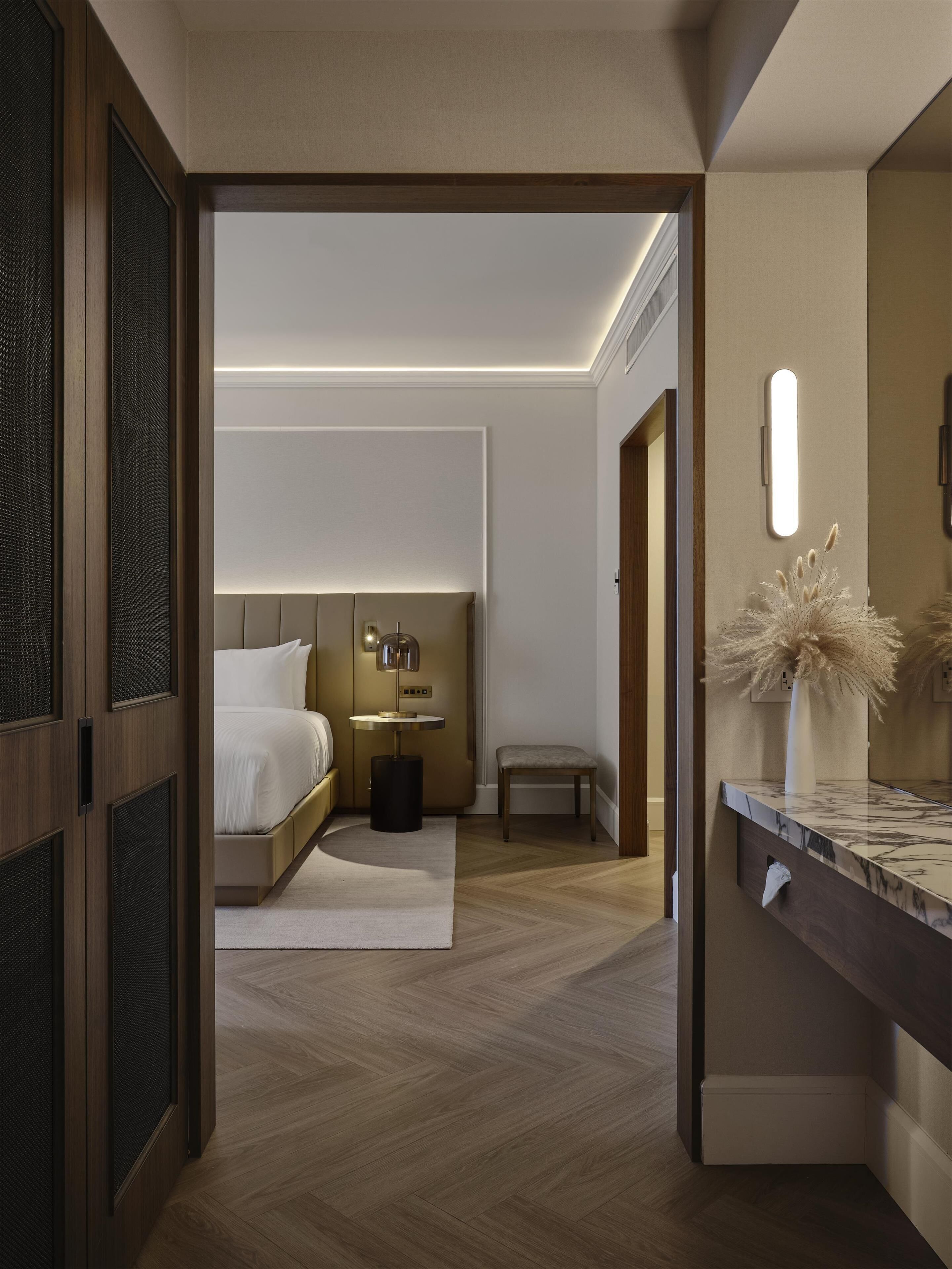 Een moderne hotelkamer met een houten vloer, een bed met witte lakens, een nachtkastje met een lamp en een klein bankje.