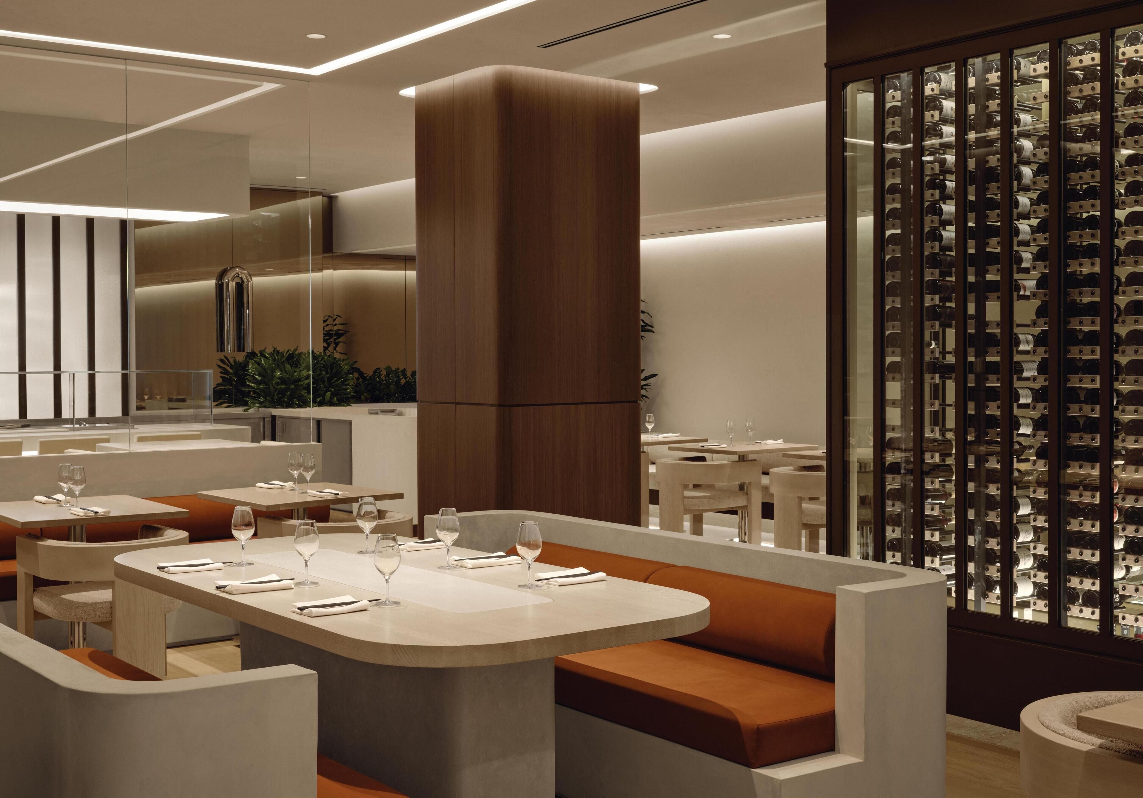 Un restaurante moderno con un espacio central de asientos que incluye una gran mesa redonda y bancos acolchados color naranja. Se observan varias mesas de comedor con manteles blancos y copas de vino dispuestas. La decoración incluye una columna de madera grande y un panel de vidrio con diseño ornamental.