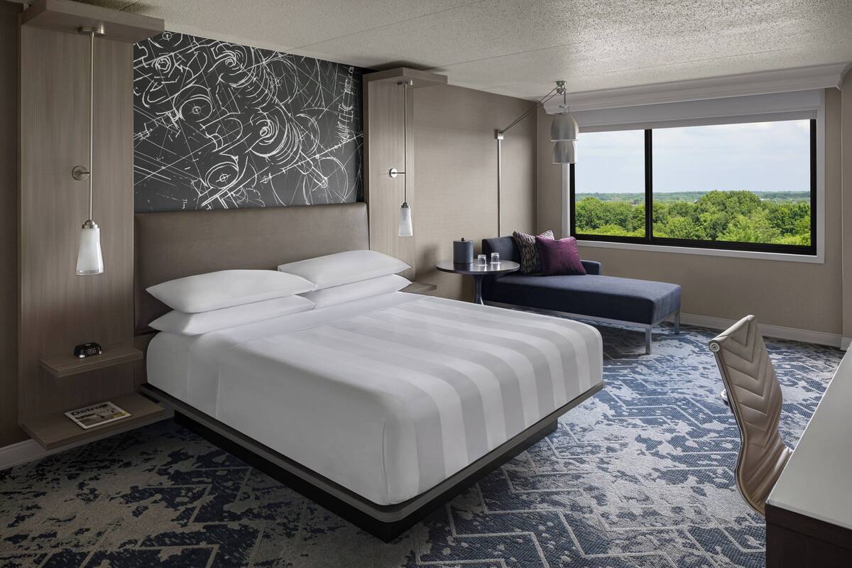 Une chambre d’hôtel moderne, dotée d’un grand lit, de tables de chevet, d’un petit bureau et d’une fenêtre donnant sur un paysage verdoyant.